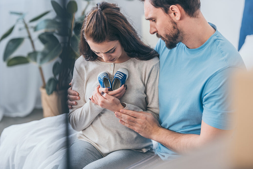 selective focus of sad husband hugging grieving wife with baby shoes